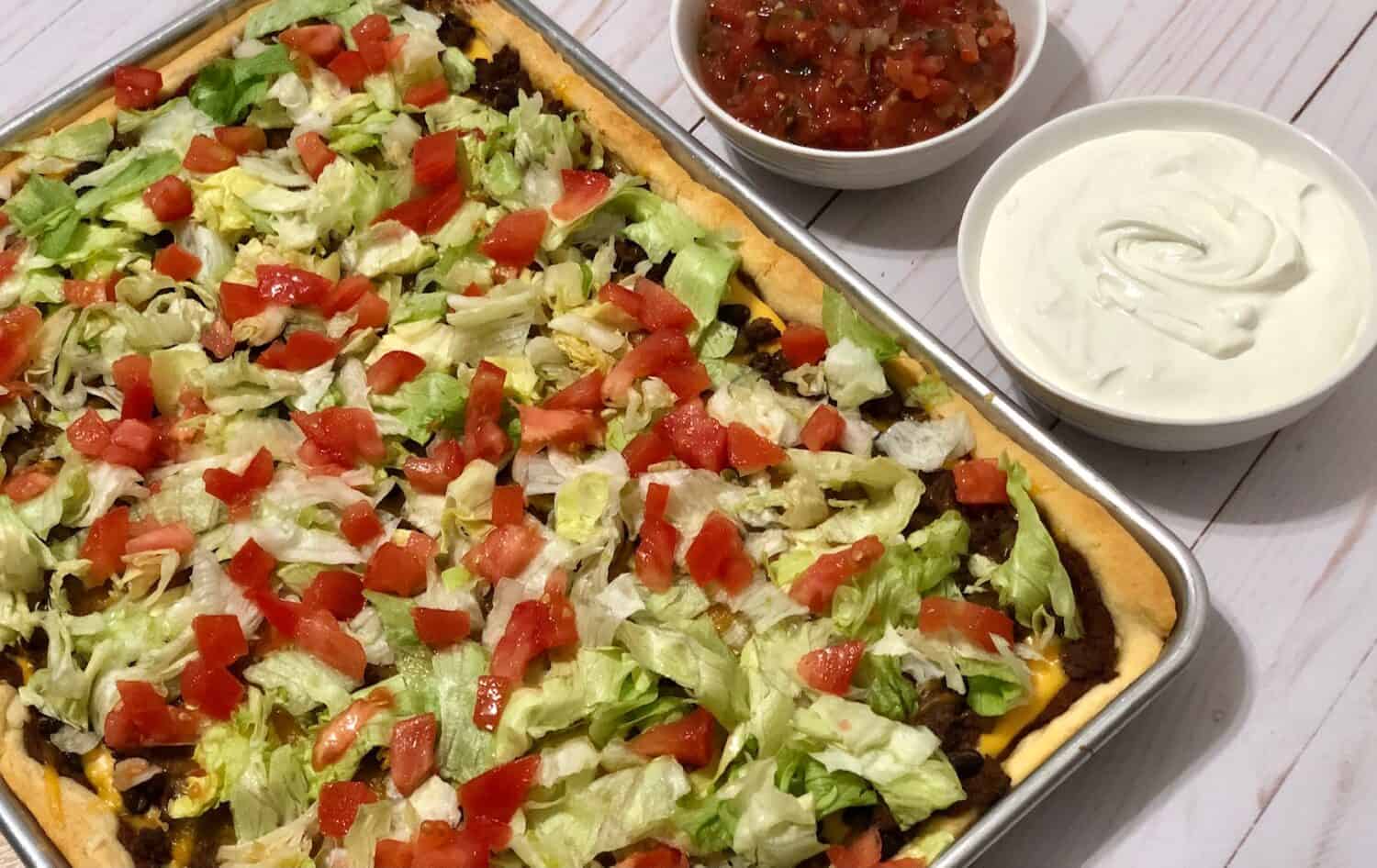 A golden, crescent roll sheet pan taco pizza loaded with seasoned ground beef, melted cheese, refried beans, black or pinto beans, jalapeños, fresh tomatoes and lettuce Served on a baking sheet with a side of sour cream and salsa.