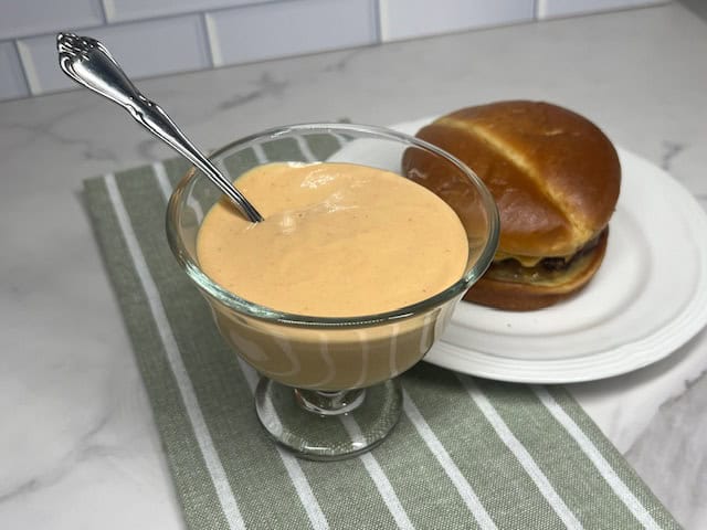 A small bowl of creamy homemade burger sauce with a spoon and a burger.