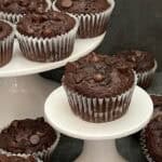 A counter top. full of double chocolate muffins with a cracked top, revealing a soft center, sitting on a white plate with chocolate chips bursting out of the top.