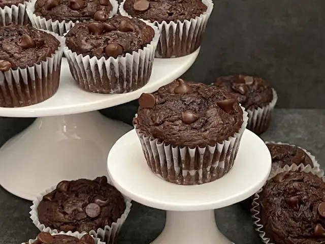 A counter top. full of double chocolate muffins with a cracked top, revealing a soft center, sitting on a white plate with chocolate chips bursting out of the top.