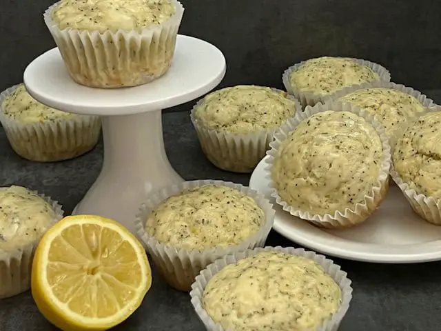 A batch of golden lemon poppy seed muffins with a light drizzle of lemon glaze, sitting on a white cake plate.