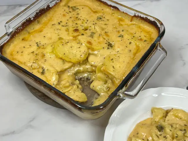 A steaming baking dish full of cheesy scalloped potatoes topped with parsley and bacon crumlbles